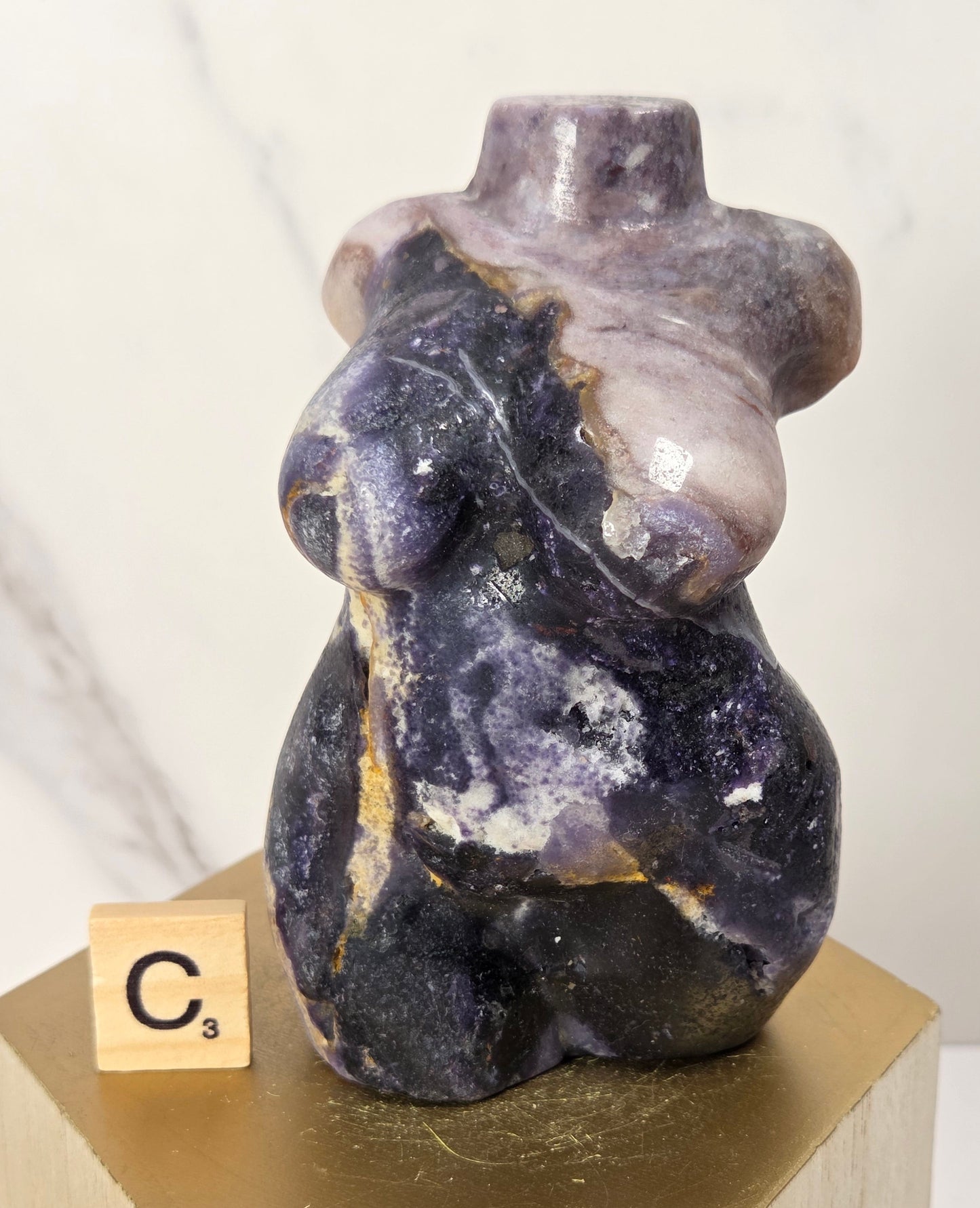 A small sculptural figurine resembling a seated female form made from polished stone, shown on a gold hexagonal pedestal with a wooden block labeled C3 beside it. The stone has a marbled mix of deep purple, gray, black, and pale lavender tones.