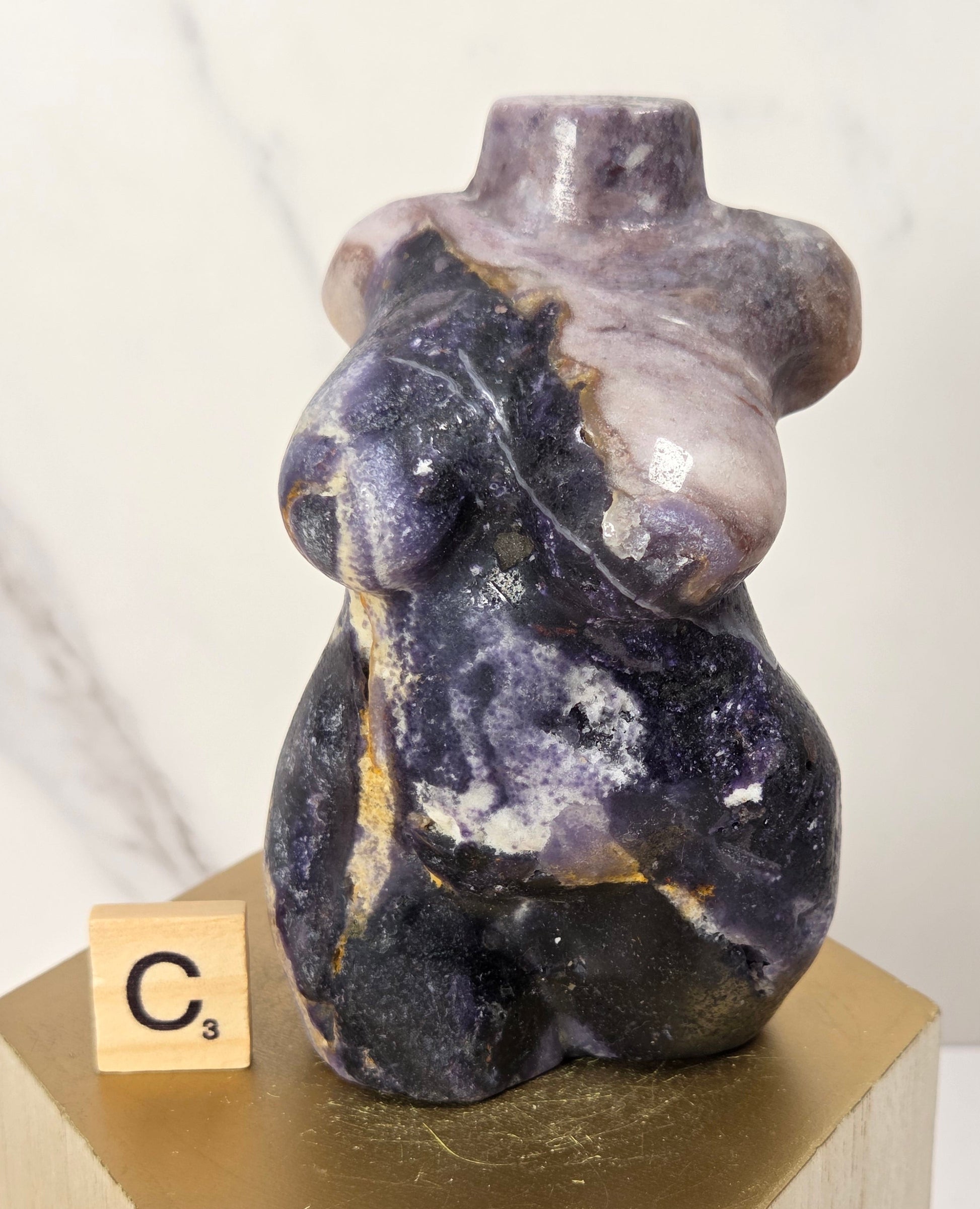 A small sculptural figurine resembling a seated female form made from polished stone, shown on a gold hexagonal pedestal with a wooden block labeled C3 beside it. The stone has a marbled mix of deep purple, gray, black, and pale lavender tones.