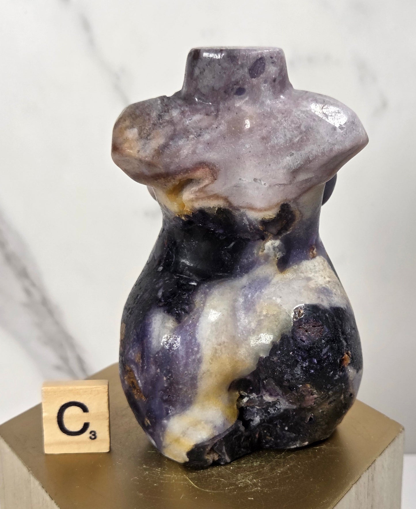 A small sculptural vase or vessel made from marbled purple, cream, and dark stone, displayed on a gold hexagonal pedestal with a wooden block labeled C3 in front, against a light marble-pattern background.
