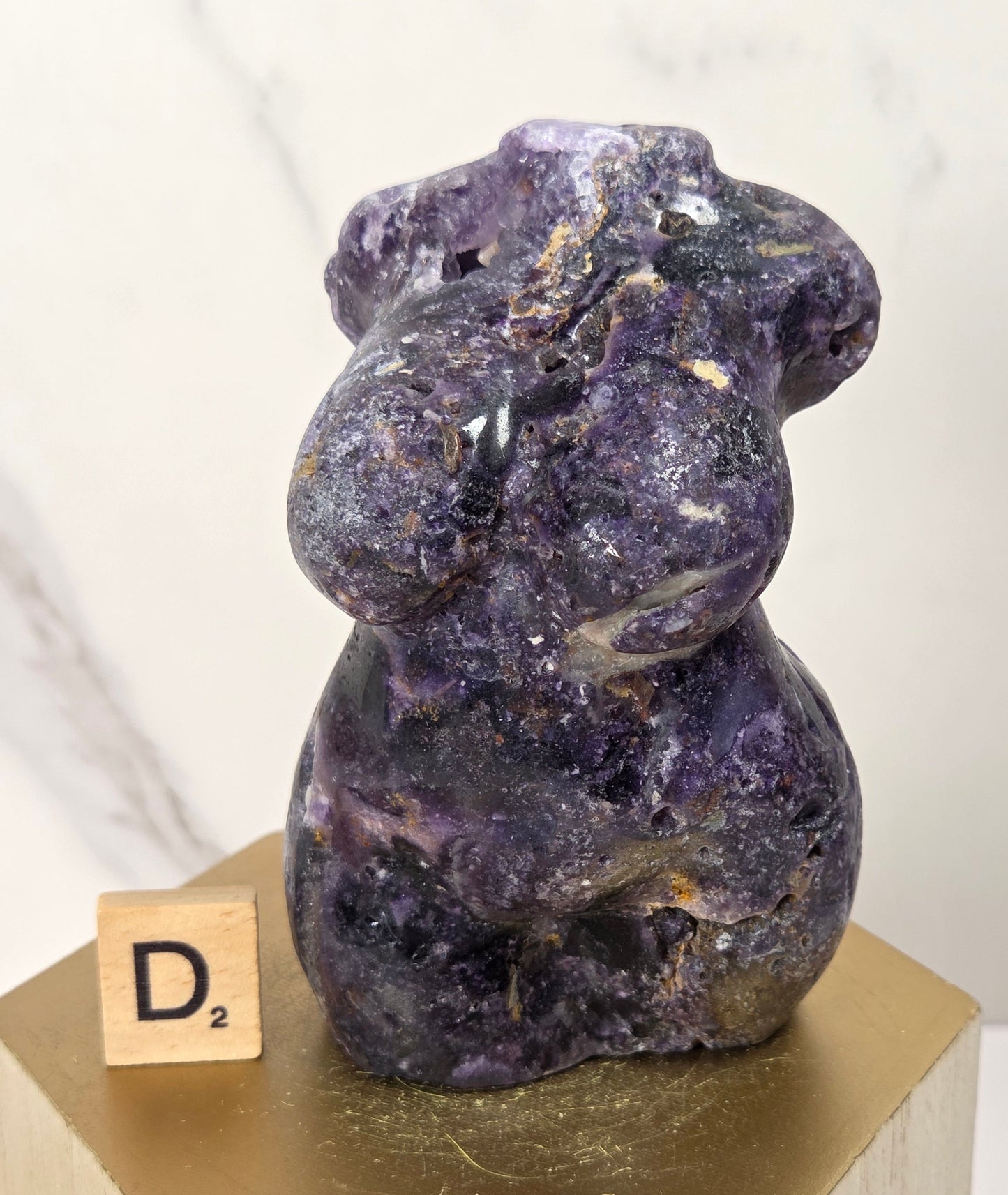 A small carved purple stone figurine with a rough, organic texture sits on a gold hexagonal display block next to a wooden Scrabble tile showing the letter 'D'. The background is a clean, light marble-patterned wall.
