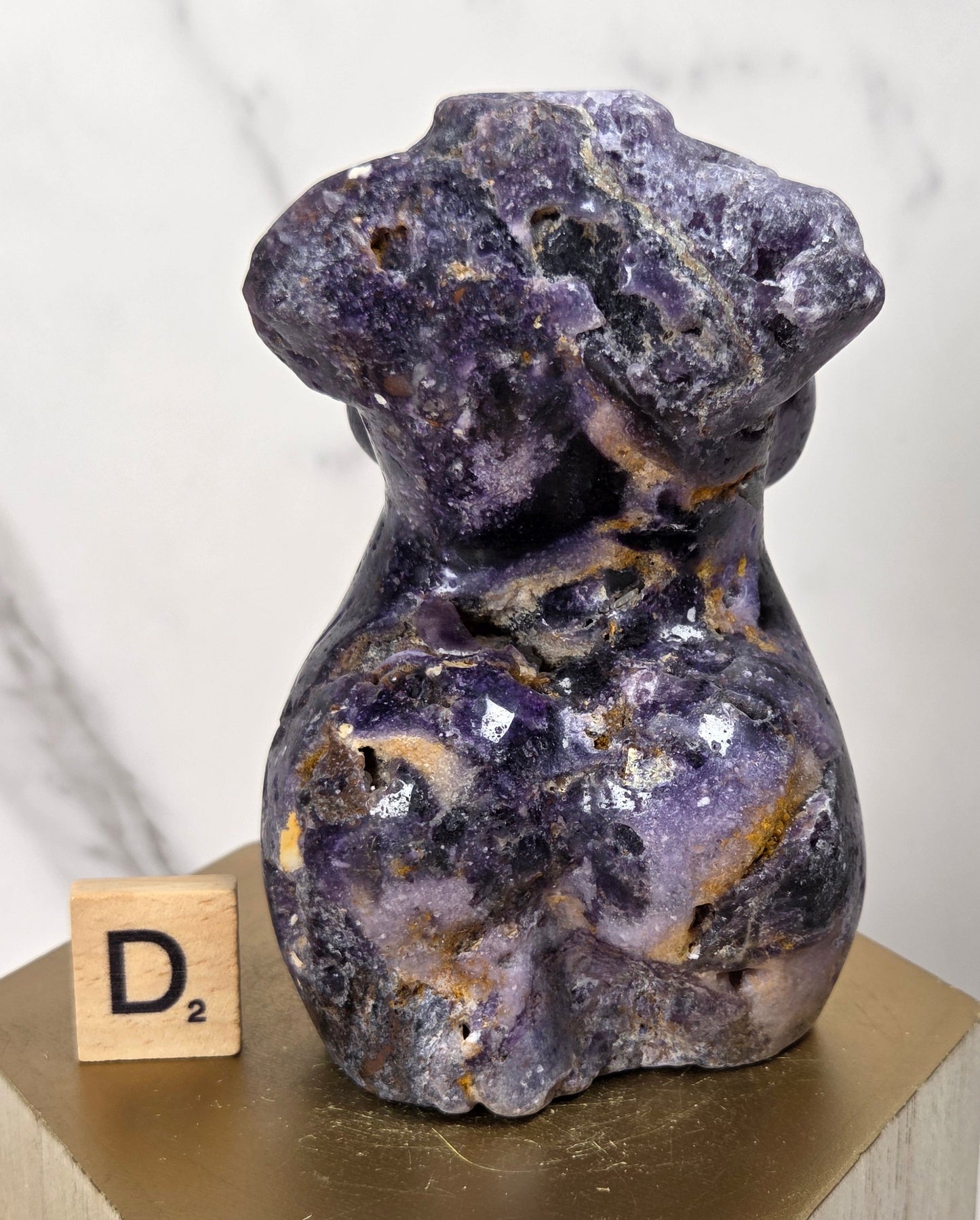 A small sculpted purple stone figurine with natural marbling and crystalline texture, displayed on a gold-toned pedestal next to a wooden Scrabble tile showing D2, against a light marble-patterned background.