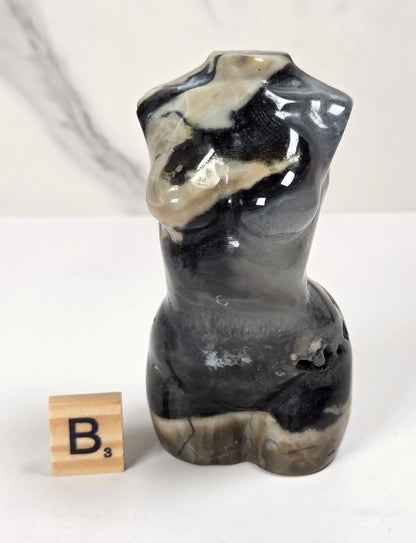 A small sculptural torso figurine made from polished black, gray, and beige agate or marble, standing on a white surface next to a wooden Scrabble tile showing the letter 'B'.