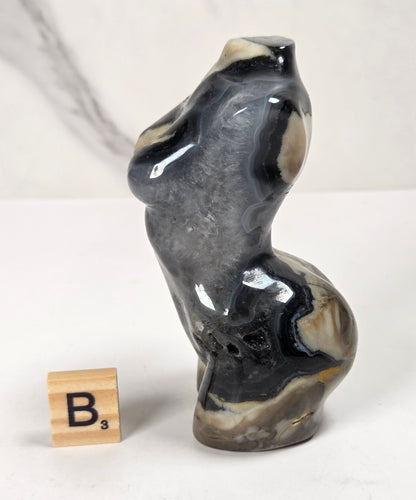 A small sculptural figurine resembling a stylized female form, made from polished agate or marble with swirling black, gray, white, and beige bands. It is displayed on a white surface next to a wooden Scrabble tile showing the letter 'B'.