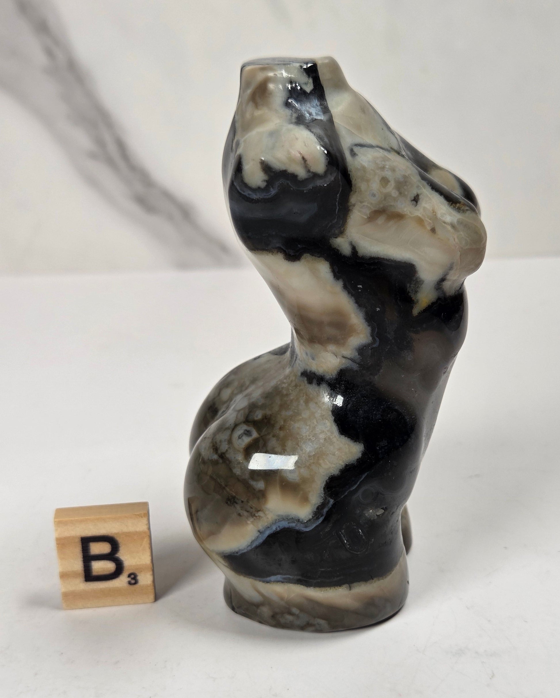 A small sculptural figurine resembling a stylized female torso with a glossy black, beige, and gray marbled stone-like finish, shown standing on a white surface next to a wooden Scrabble tile marked B3.