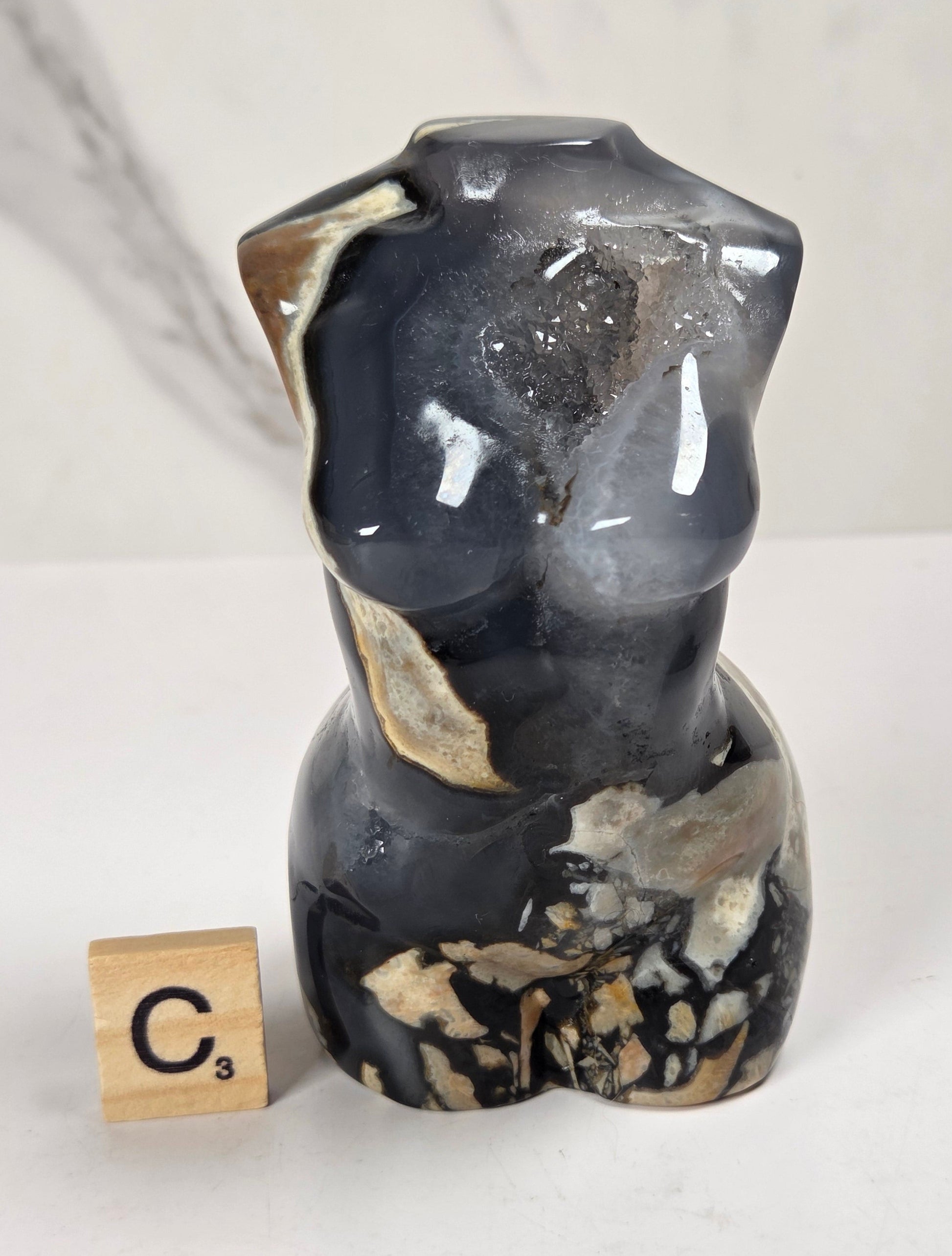 A small sculptural torso figurine made from polished black agate with natural beige and gray inclusions, displayed on a white surface next to a wooden Scrabble tile showing the letter C.