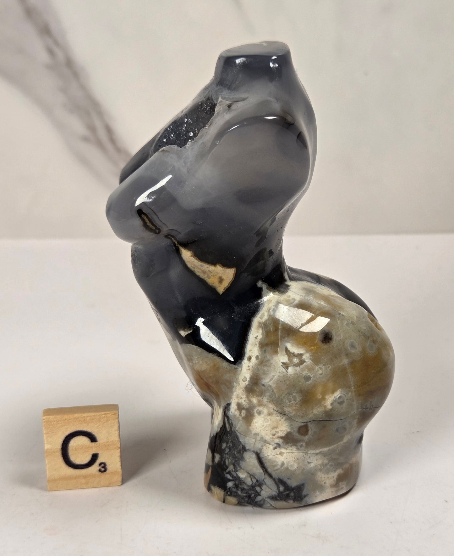 A small sculptural bust made from polished agate or marble, featuring a dark charcoal-black upper section and a mottled beige, gray, and brown lower section. It is displayed on a light surface next to a wooden Scrabble tile showing the letter 'C' with a 3-point value.