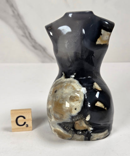 A glossy black and beige agate stone torso-shaped vase displayed on a white surface next to a wooden Scrabble tile showing the letter 'C'.