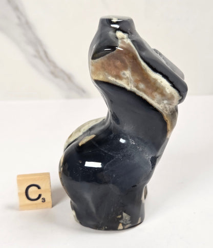 A small sculptural figurine of a badger in glossy black with natural beige and brown stone veining, shown next to a wooden Scrabble tile on a white marble-patterned surface.