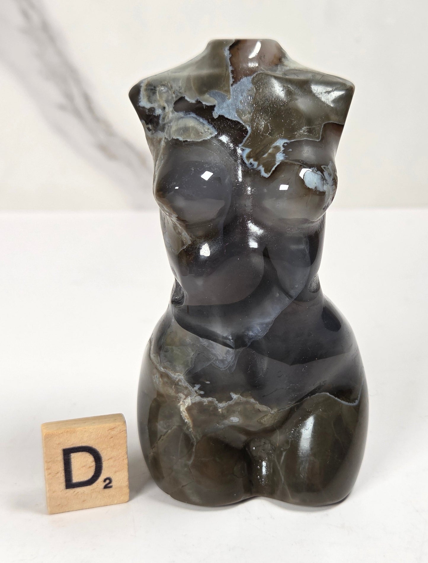 A polished dark agate bust sculpture of a female torso, displayed next to a wooden Scrabble tile on a white marble-patterned surface.