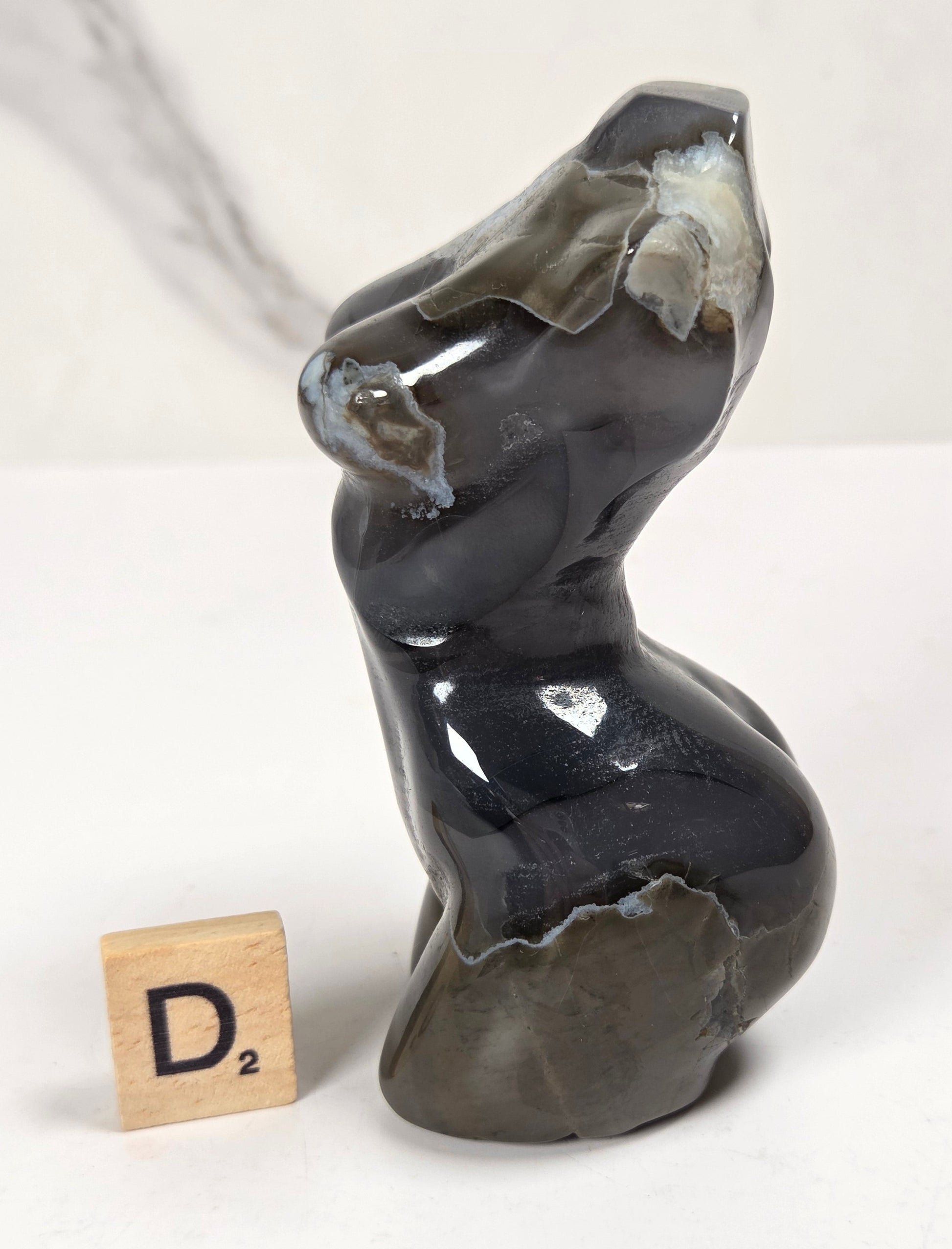 A small abstract sculptural object made from polished dark stone with natural lighter patches, shown on a white surface with a small wooden Scrabble tile marked D2 beside it.