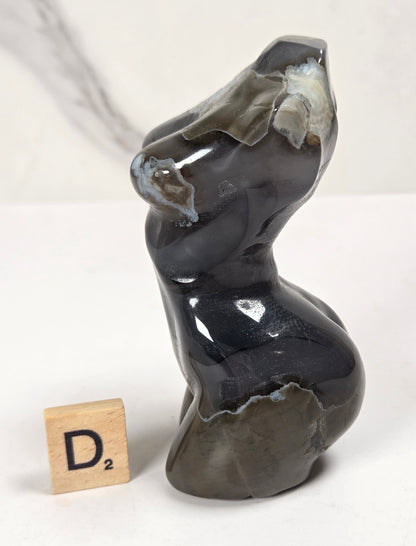 A small abstract sculptural object made from polished dark stone with natural lighter patches, shown on a white surface with a small wooden Scrabble tile marked D2 beside it.