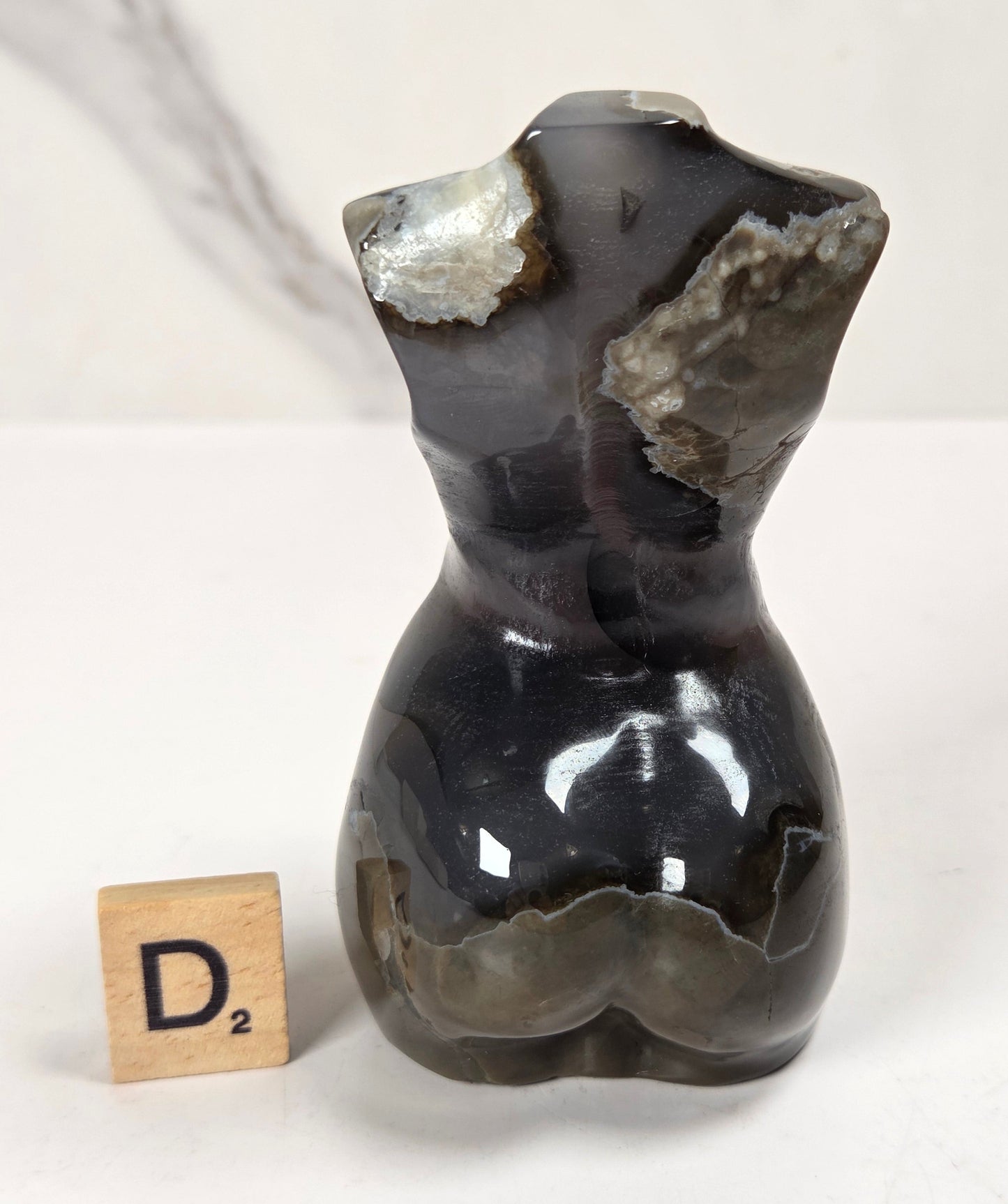 A small sculptural torso-shaped object made of polished dark agate or stone, with natural lighter-colored crystalline sections exposed near the shoulders, shown on a white surface with a small wooden letter tile beside it.