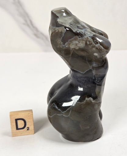 A polished dark agate figurine shaped like a stylized female torso, displayed next to a small wooden Scrabble tile on a white surface with a light gray marble-patterned background.