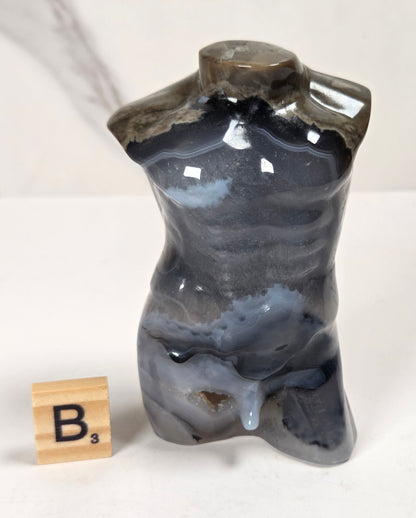 A small polished stone torso sculpture with a glossy marbled finish in dark gray, blue-gray, and brown tones, displayed on a white surface next to a wooden Scrabble tile showing the letter B.