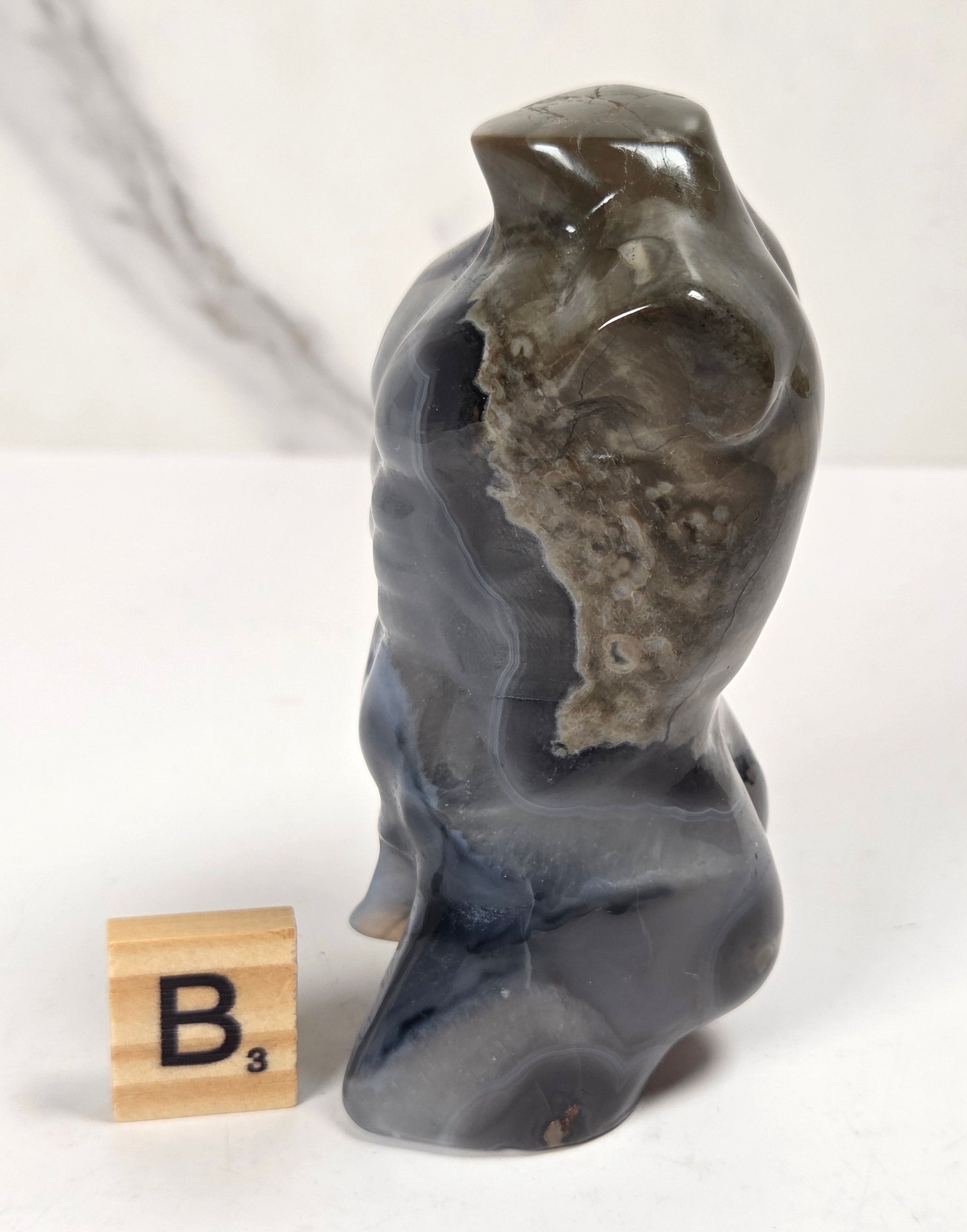 A small carved stone figurine resembling a seated figure or animal, shown on a white surface with a marble-patterned backdrop. A wooden Scrabble tile with the letter B is placed beside it for scale.