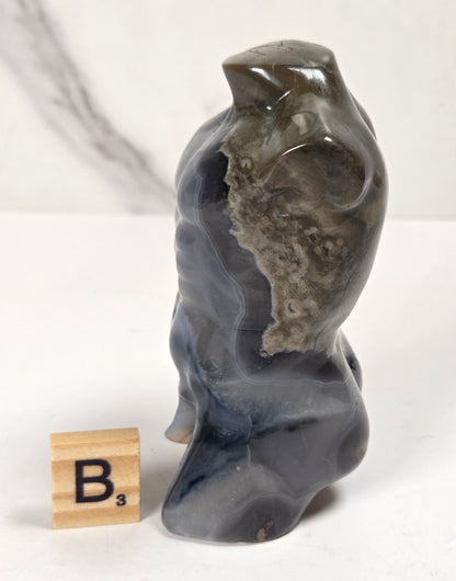 A small carved stone figurine resembling a seated figure or animal, shown on a white surface with a marble-patterned backdrop. A wooden Scrabble tile with the letter B is placed beside it for scale.