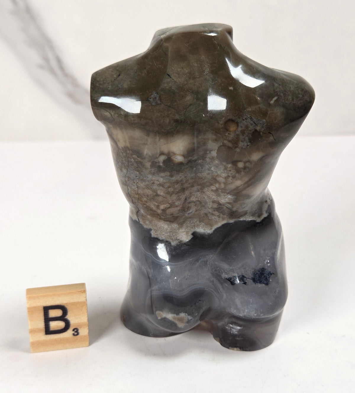A polished natural stone male torso sculpture with a glossy finish, displayed next to a wooden Scrabble tile showing the letter 'B'. The background is a clean white surface with a light gray marble-patterned wall.