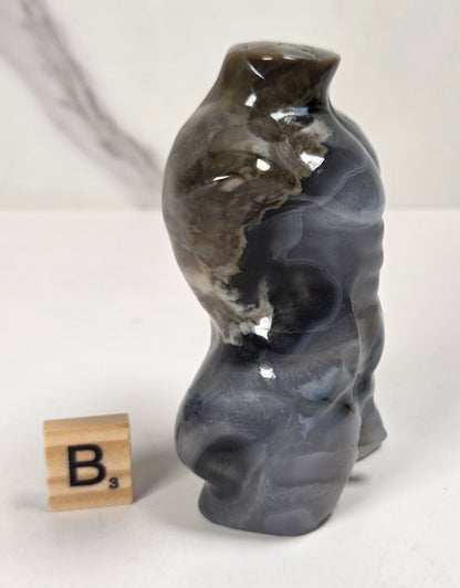 A small sculptural vase or bottle with a glossy marbled finish in dark gray, black, and brown tones, standing on a white surface next to a wooden Scrabble tile showing the letter B.