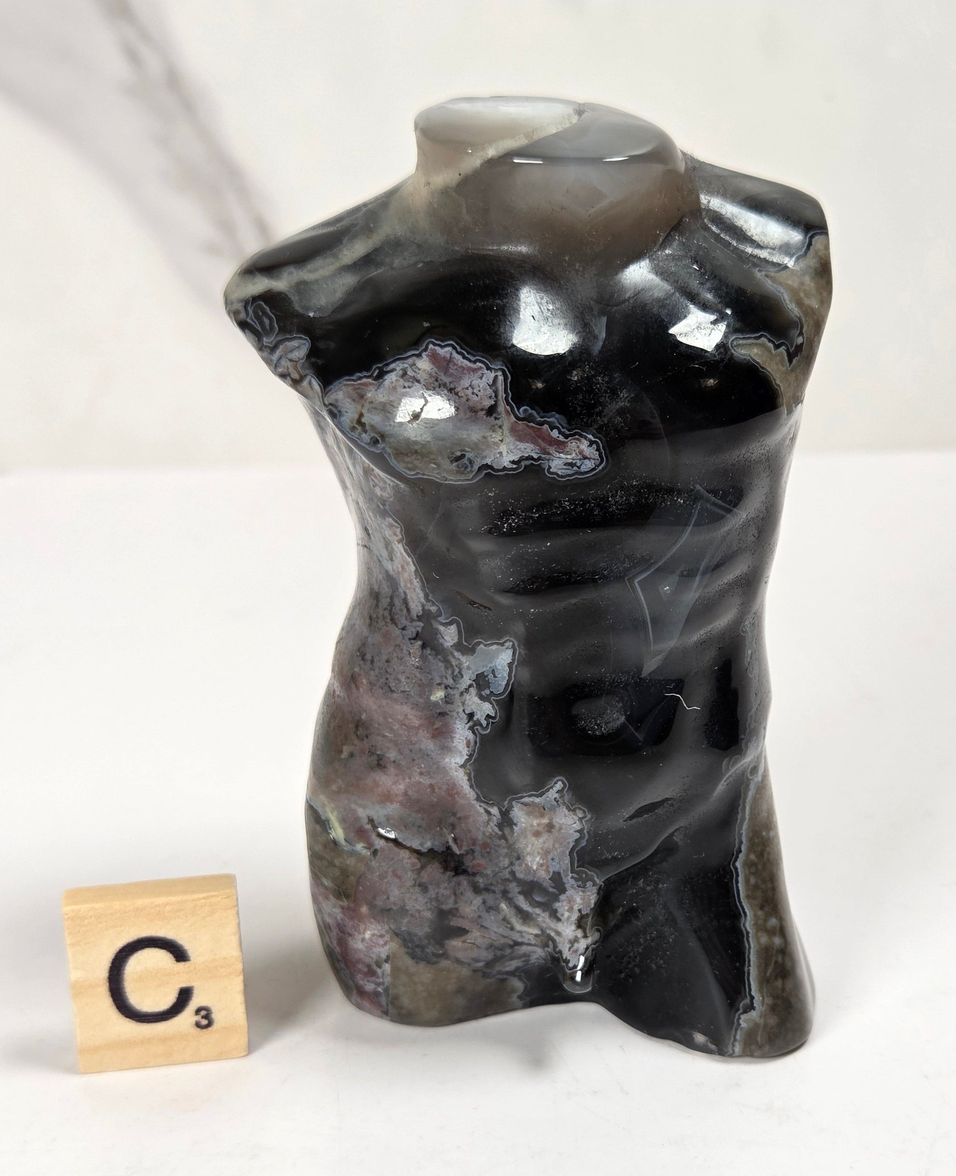 A small polished stone torso sculpture with a glossy black and gray agate-like surface, shown on a white surface next to a wooden Scrabble tile marked C3.