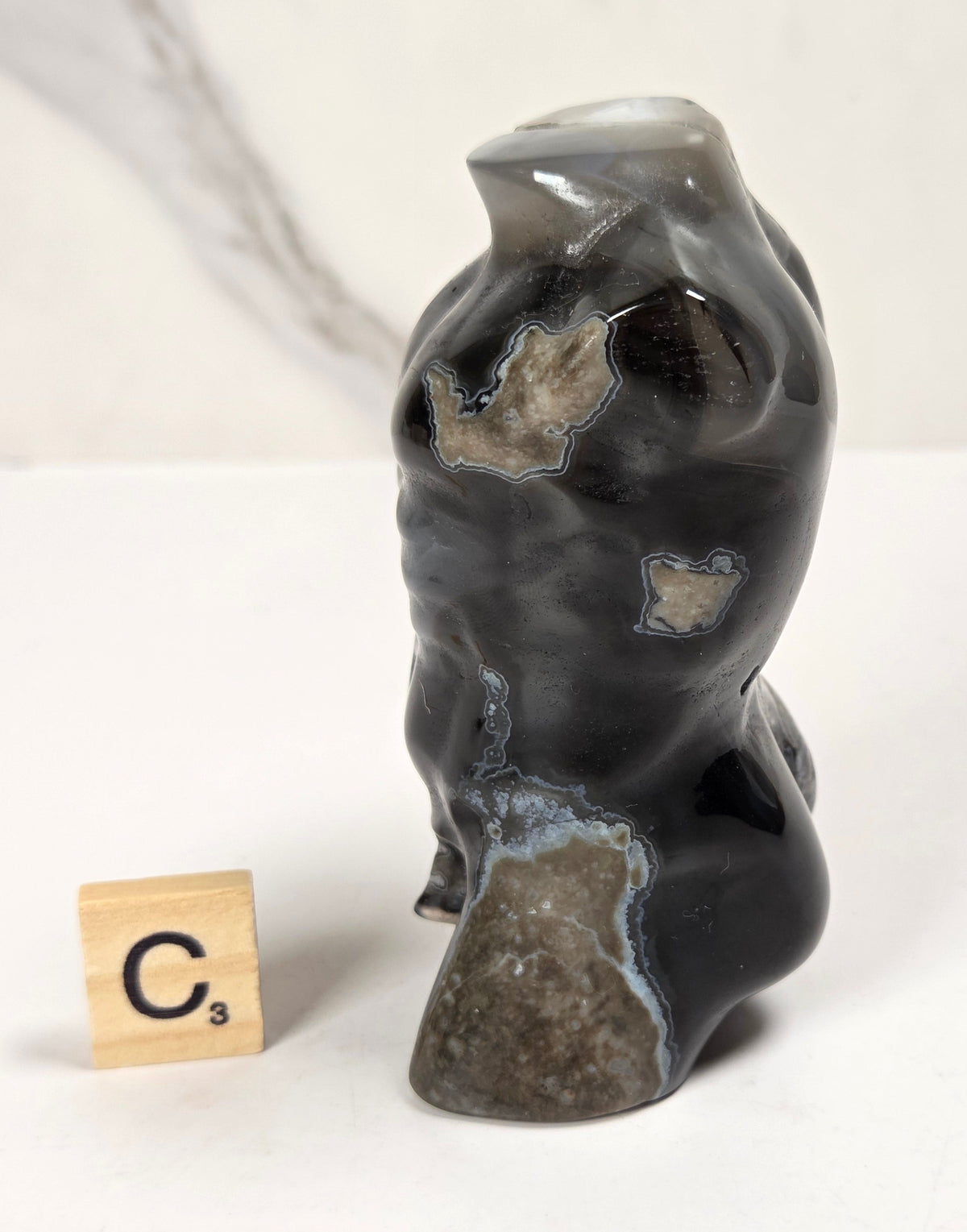 A polished black agate stone figurine shaped like a stylized torso, shown next to a wooden Scrabble tile on a white surface with a light marble background.