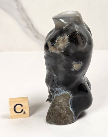 A polished black agate stone figurine shaped like a stylized torso, shown next to a wooden Scrabble tile on a white surface with a light marble background.