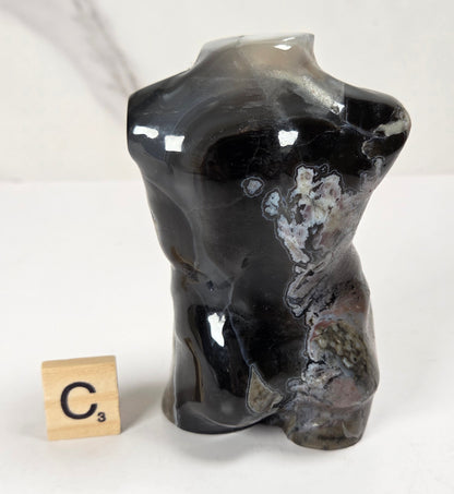 A glossy black and gray agate stone torso sculpture with natural swirling patterns, displayed next to a wooden Scrabble tile on a white marble-patterned surface.