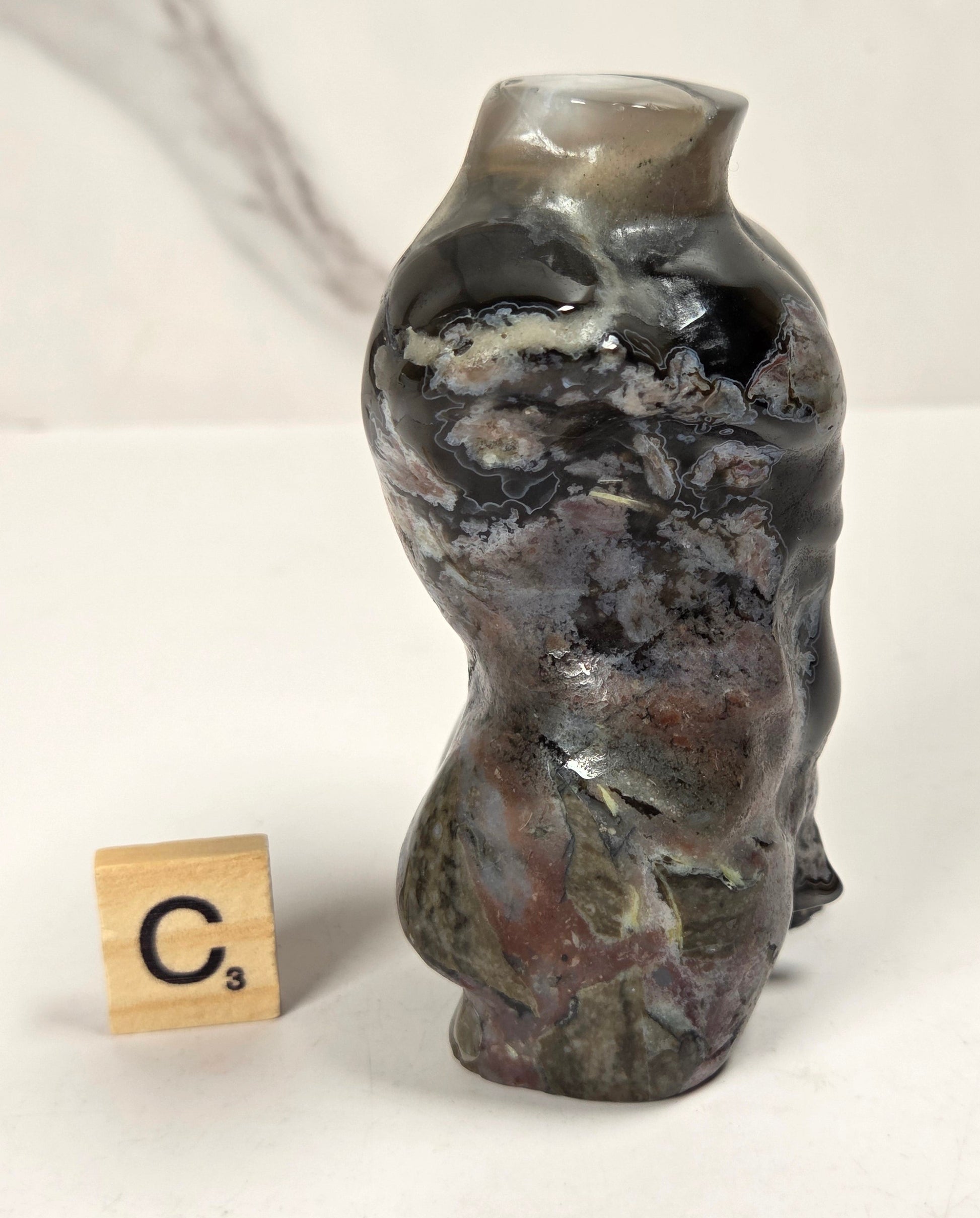 A small sculptural ceramic vase with an organic, wavy silhouette and a glossy marbled glaze in dark gray, black, brown, and muted green tones. A small wooden Scrabble tile stands beside it on a white surface against a light marble-patterned background.