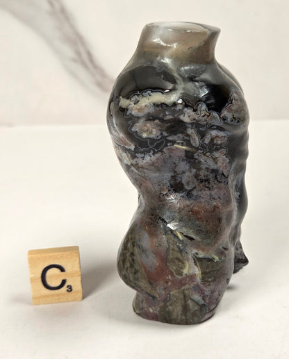 A small sculptural ceramic vase with an organic, wavy silhouette and a glossy marbled glaze in dark gray, black, brown, and muted green tones. A small wooden Scrabble tile stands beside it on a white surface against a light marble-patterned background.