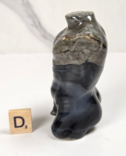 A polished natural stone figurine with a layered appearance, featuring a dark black lower section and a gray-brown upper section with natural inclusions, shown next to a small wooden Scrabble tile on a white marble-patterned surface.
