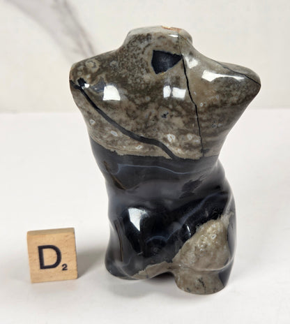 A small polished stone figurine with a split or fractured top section, shown on a white surface with a small wooden letter block beside it. The background is a light marble-patterned wall.