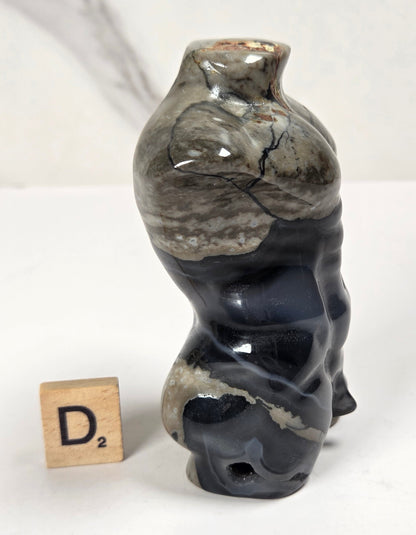 A small sculptural ceramic vase with an organic, wavy silhouette in glossy black and gray marbled glaze, shown on a white surface next to a wooden Scrabble tile.