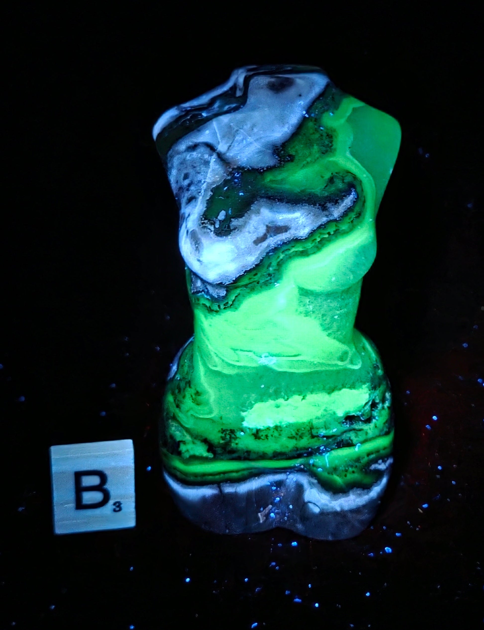A small sculptural object with a marbled green, black, and white surface, photographed on a black background. A small white square label with the letters 'B3' appears near the lower left.