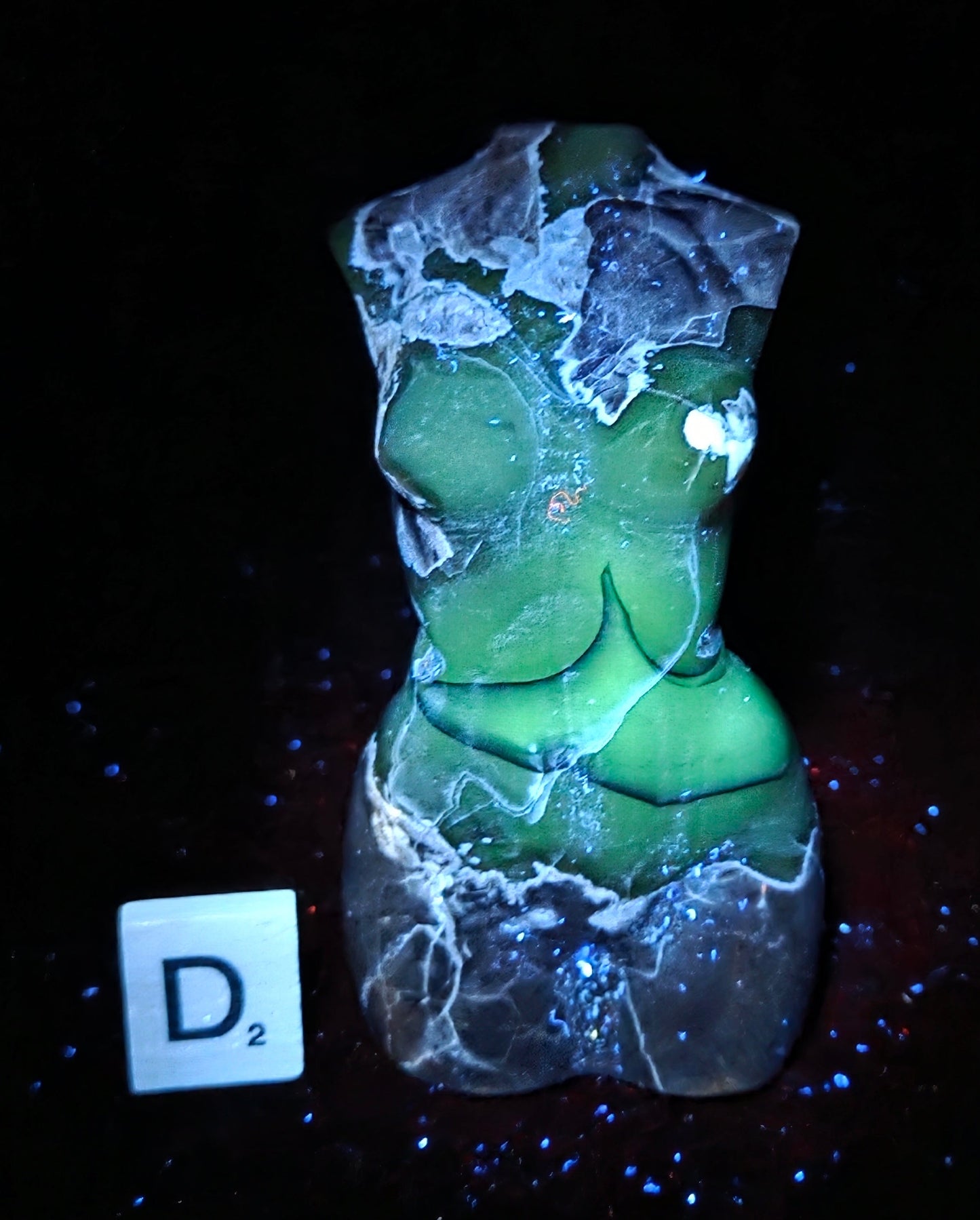 A small sculptural bust or torso-shaped object with a cracked, stone-like surface, shown glowing green under UV light on a black background. A small white tile with the letter D2 appears at lower left.
