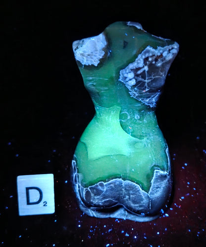 A small sculptural object with a glossy green and translucent body and rough white-gray edges, photographed on a black background with a small white tile marked D2 beside it.