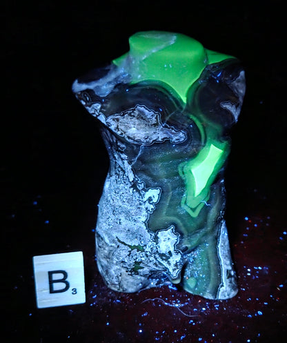 A raw, rough stone or agate slab with striking neon green, black, and gray banding, photographed under UV light on a black background with a small letter tile showing 'B3' nearby.