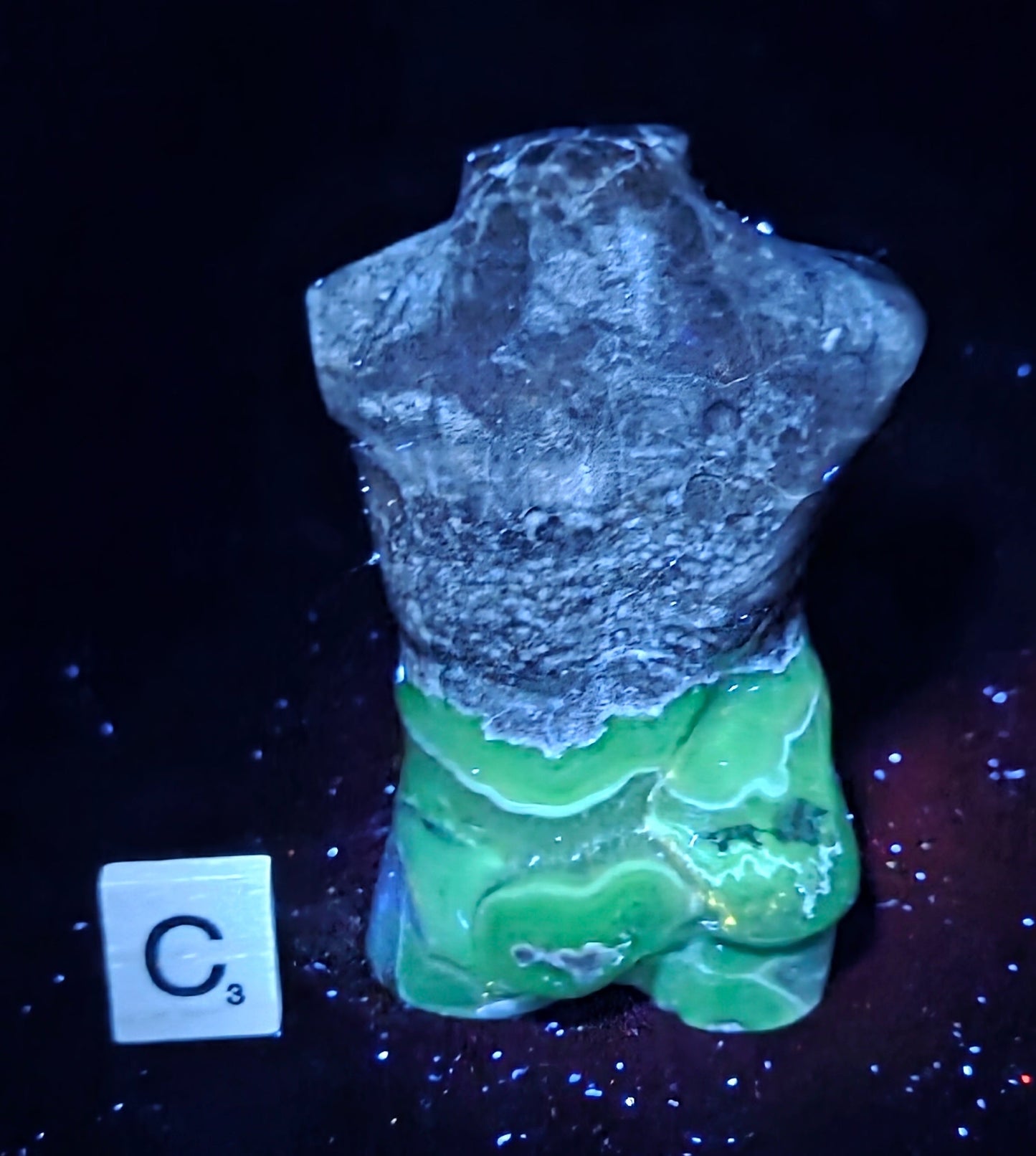 A small torso-shaped decorative object or figurine with a rough gray upper section and a bright green lower section, photographed on a dark background with a small white tile marked C3 nearby.