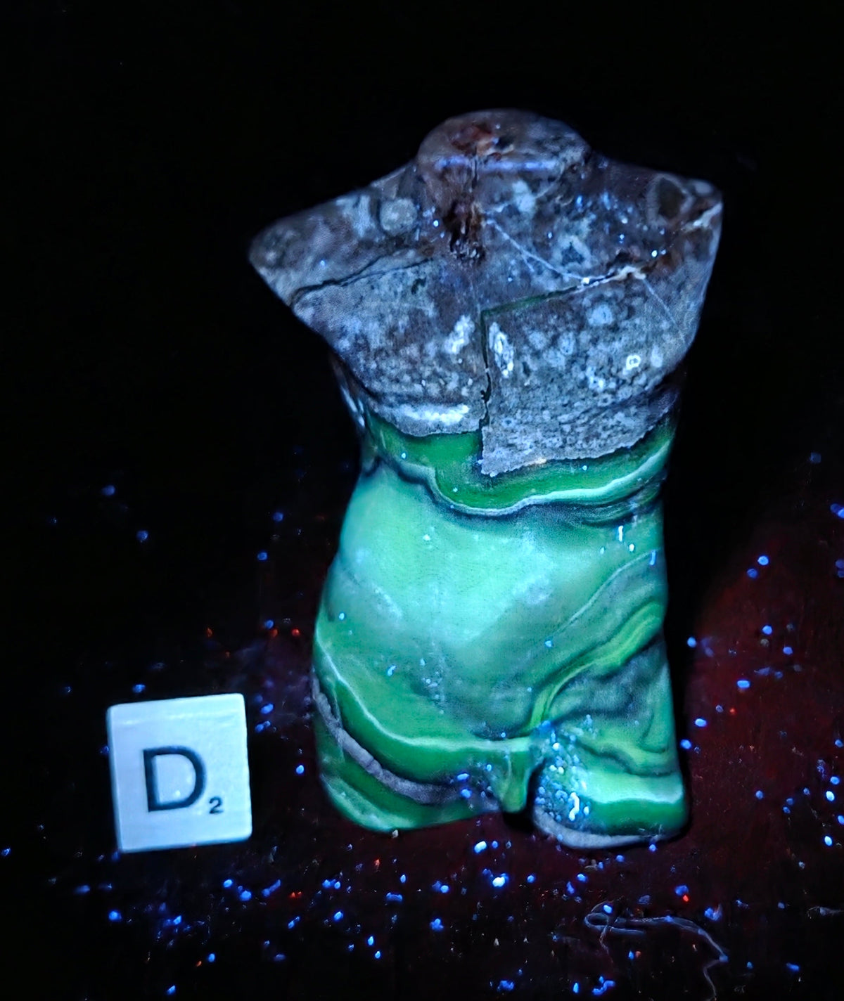 A small decorative object shaped like a torso, featuring a mottled gray top and green marbled lower half, photographed on a black background with a small white card marked "D3" beside it.