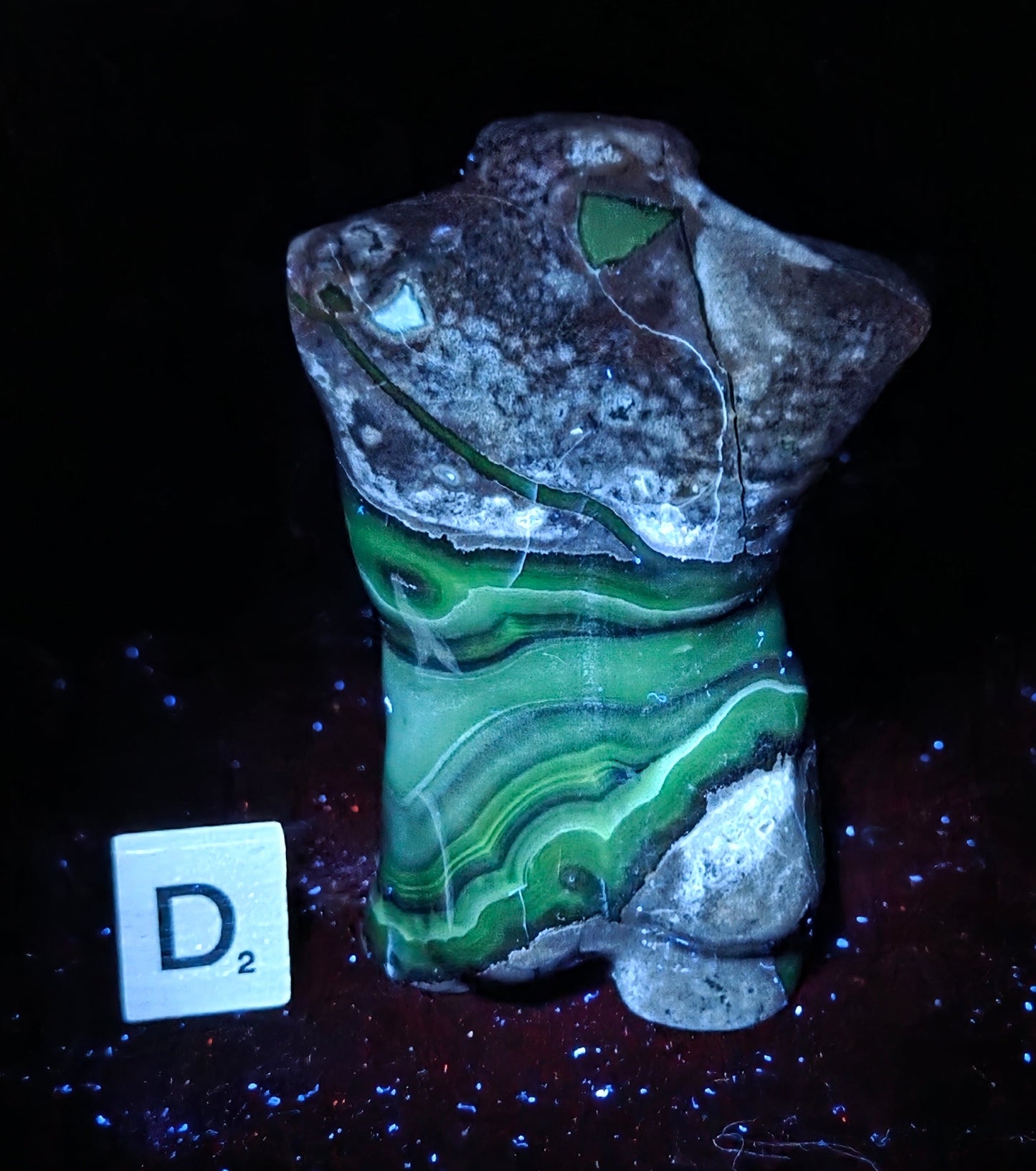 A carved stone figurine resembling a stylized torso or animal head, featuring layered green agate-like bands over a gray and white marbled base, photographed on a black background with a small white tile marked D2 beside it.