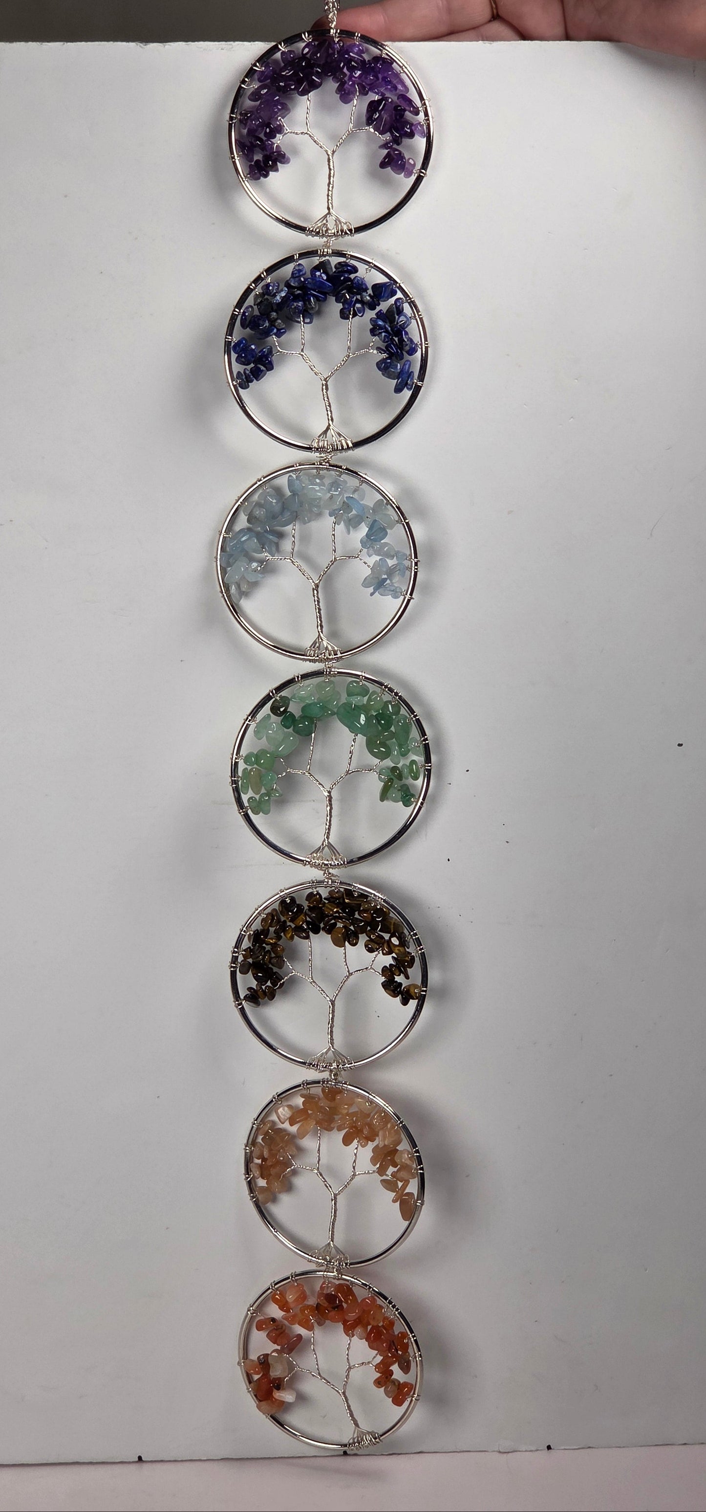 A vertical hanging suncatcher or wall hanging made of seven stacked circular wire frames with tree-of-life motifs and colored stone or resin inlays, shown against a white board with a hand holding the top.