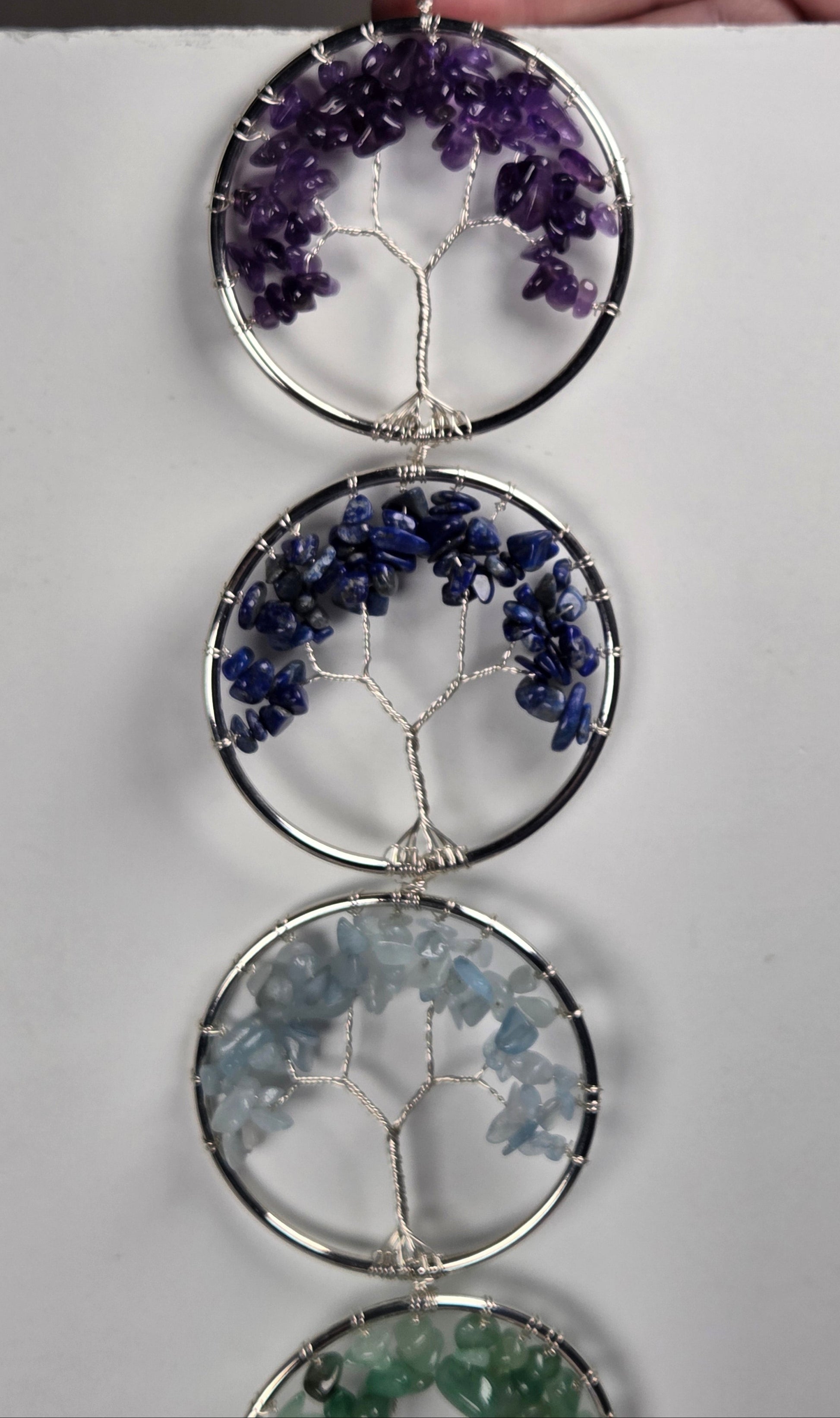 A vertical hanging wall ornament made of four circular silver frames, each containing a tree-of-life wire design filled with small gemstone chips in purple, blue, light blue, and green, shown against a plain white background.