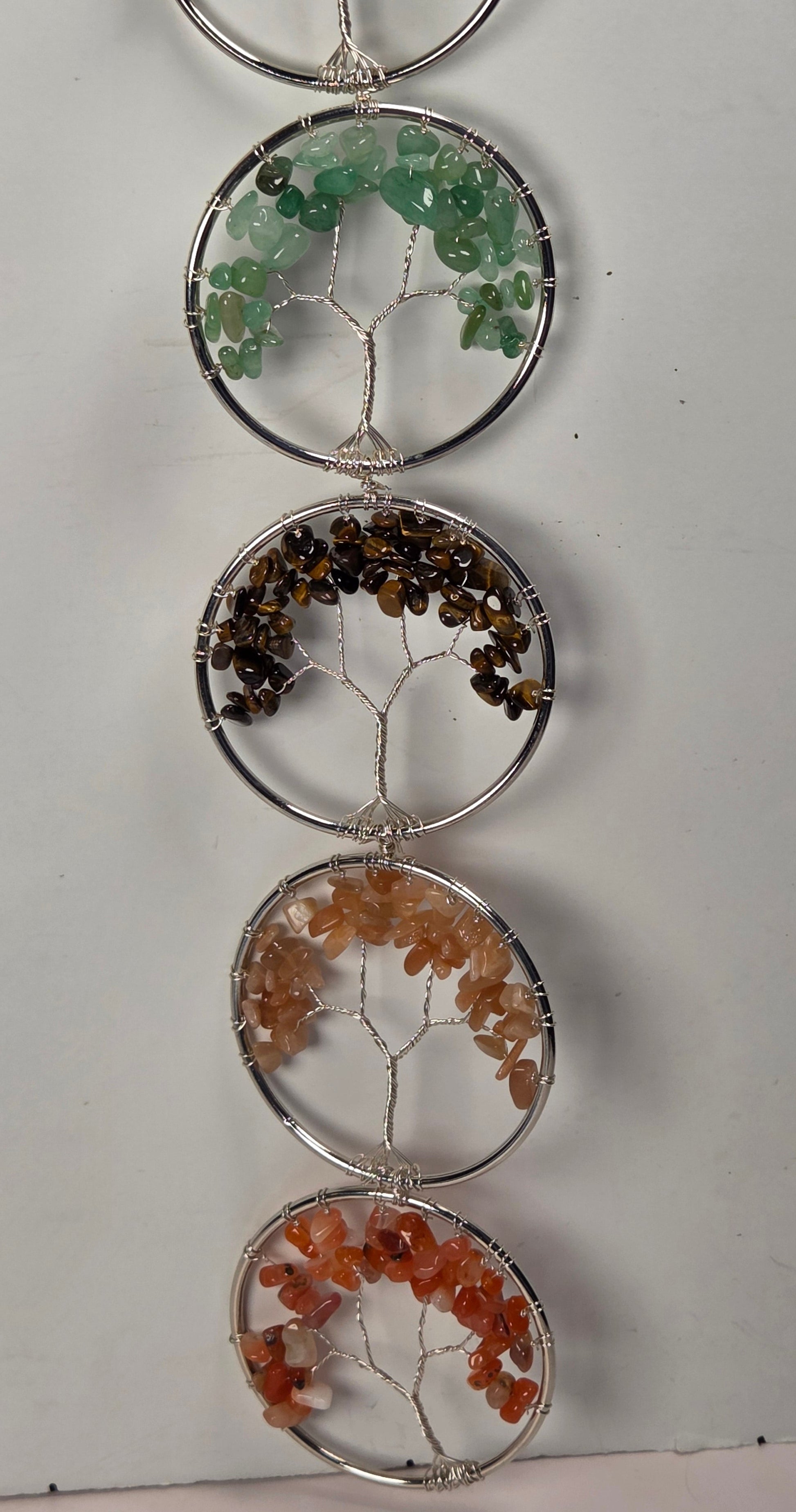 A vertical hanging crystal tree ornament made of five circular silver wire frames, each holding different colored gemstone chips. The stones include green, brown, peach, and orange, set against a plain off-white background.