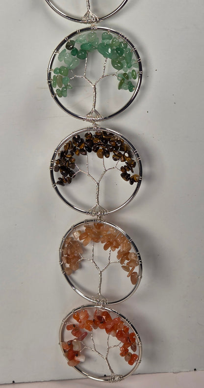 A vertical hanging crystal tree ornament made of five circular silver wire frames, each holding different colored gemstone chips. The stones include green, brown, peach, and orange, set against a plain off-white background.