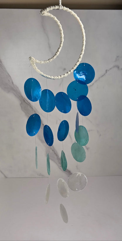 A hanging crescent moon wind chime with translucent blue and teal circular discs and white accents, suspended in front of a white marble-patterned backdrop.