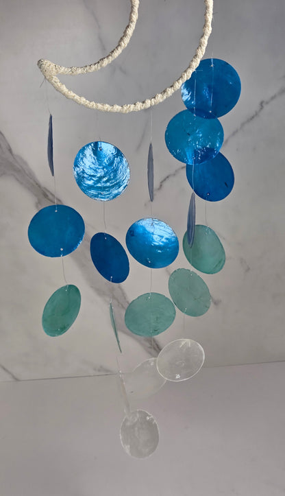 A hanging mobile with a white rope-wrapped crescent moon at the top, decorated with shiny blue and turquoise circular discs and a few clear glass discs, shown against a light marble-patterned wall.