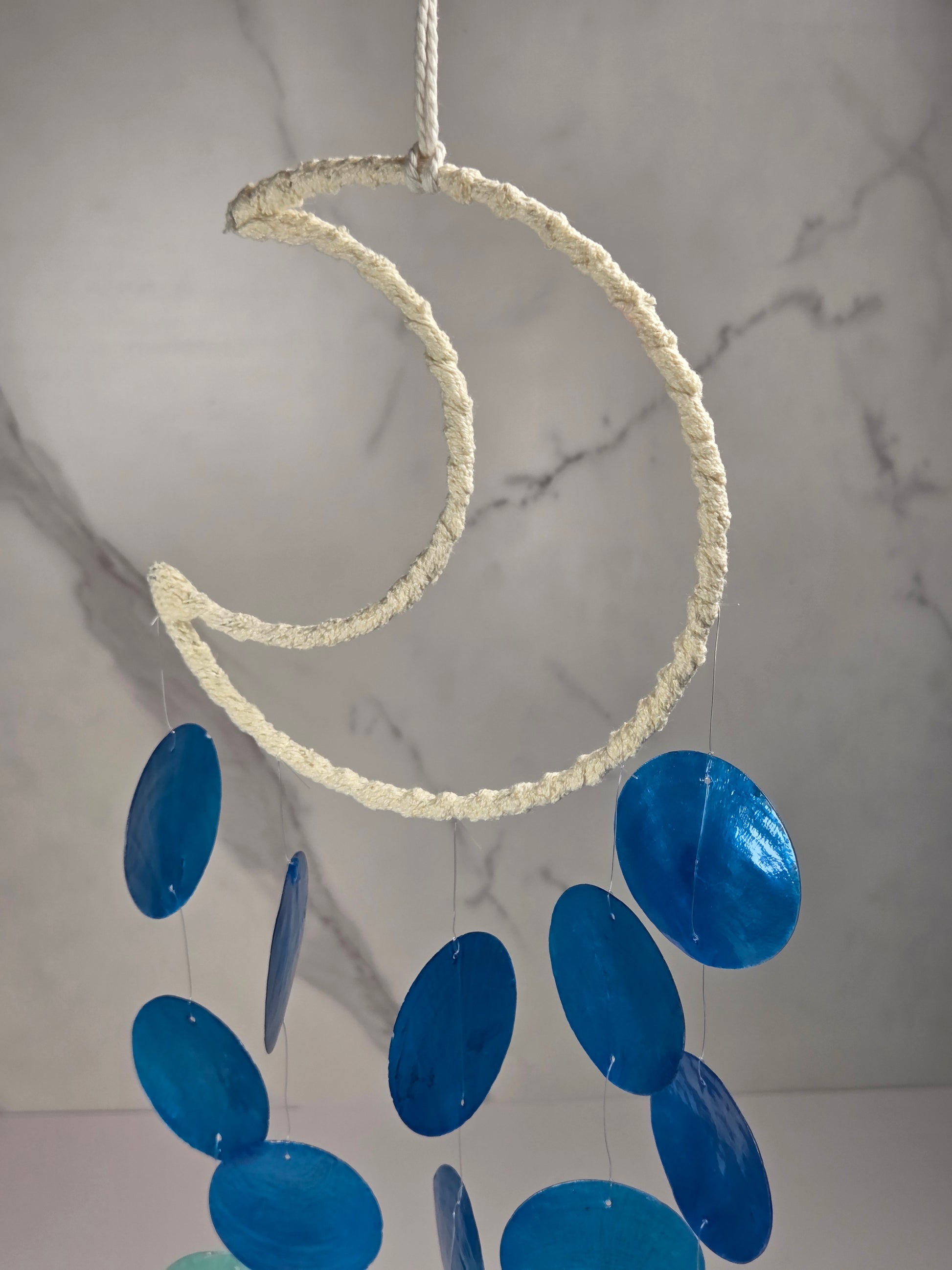 A hanging decorative mobile with a large cream rope crescent moon shape and multiple glossy blue oval discs suspended below, shown against a light marble-pattern wall.