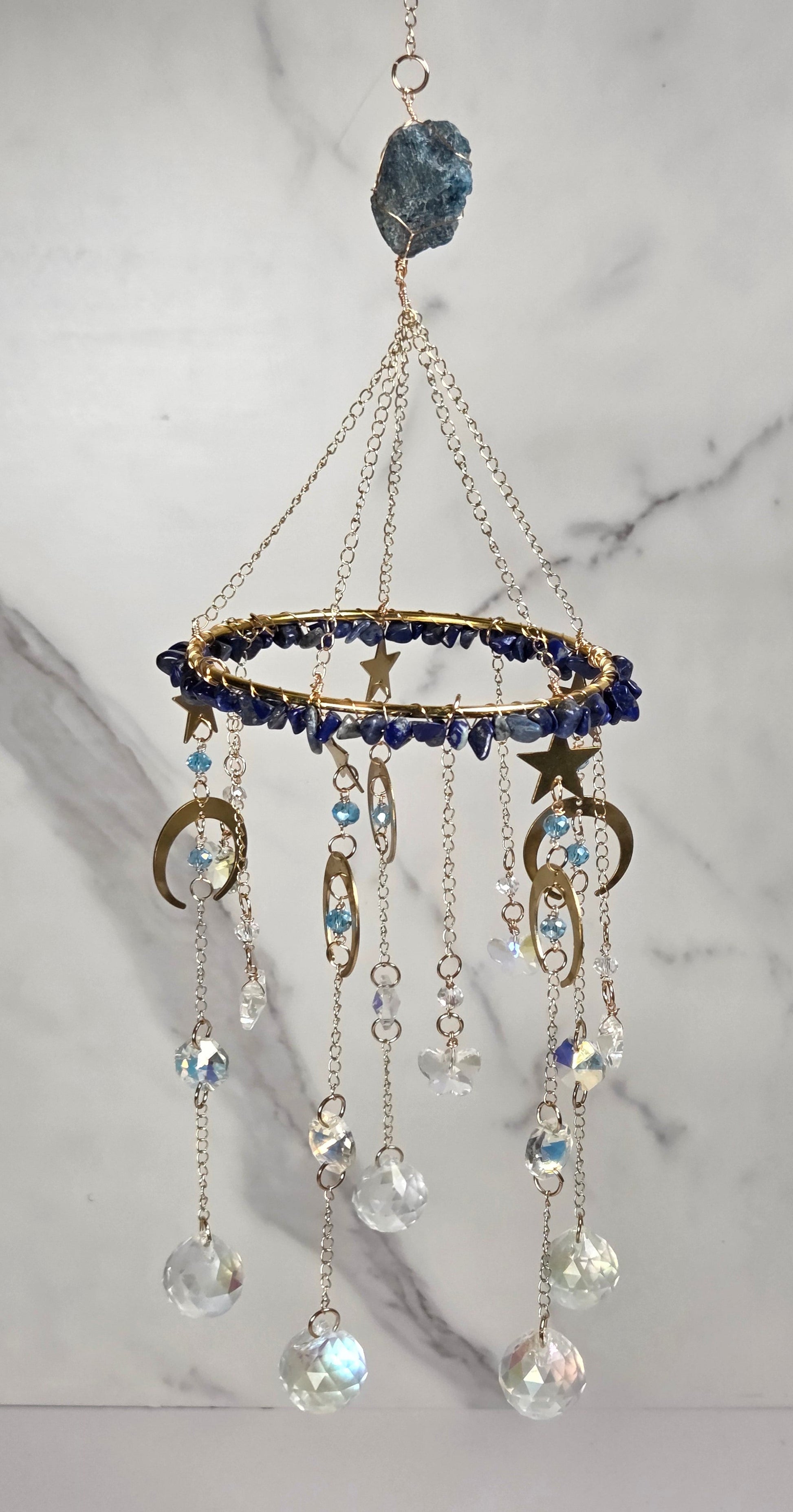 A celestial-themed mobile hanging from a gold chain, decorated with a blue crystal cluster at the top, a gold ring studded with dark blue beads, star and crescent moon charms, and dangling clear faceted beads.