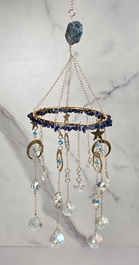 A celestial-themed mobile hanging from a gold chain, decorated with a blue crystal cluster at the top, a gold ring studded with dark blue beads, star and crescent moon charms, and dangling clear faceted beads.
