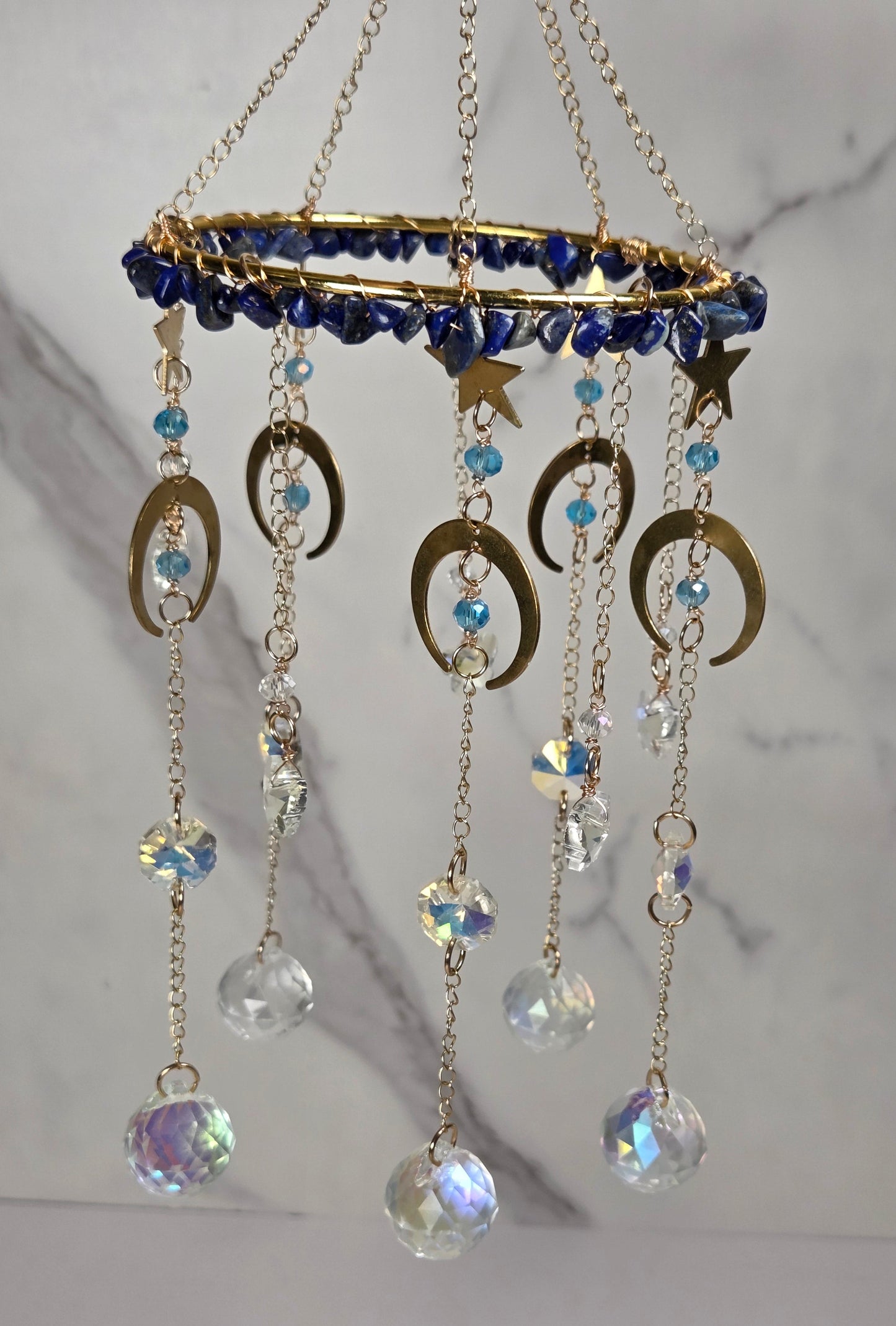 A gold-toned hanging celestial ornament with a circular top, blue beads, crescent moon and star charms, and dangling clear and iridescent crystal beads against a light marble background.