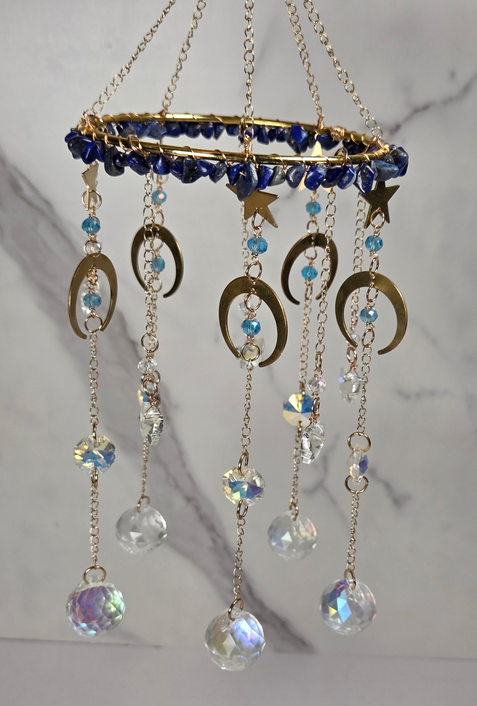 A gold-toned hanging celestial ornament with a circular top, blue beads, crescent moon and star charms, and dangling clear and iridescent crystal beads against a light marble background.