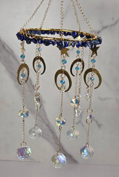 A gold-toned hanging celestial ornament with a circular top, blue beads, crescent moon and star charms, and dangling clear and iridescent crystal beads against a light marble background.